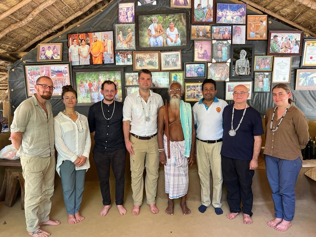 Acht Personen posieren vor einer Wand mit vielen Fotografien. Alle sind barfuss. In der Mitte ein Vertreter der Gemeinschaft der Veddah, Sri Lanka. 