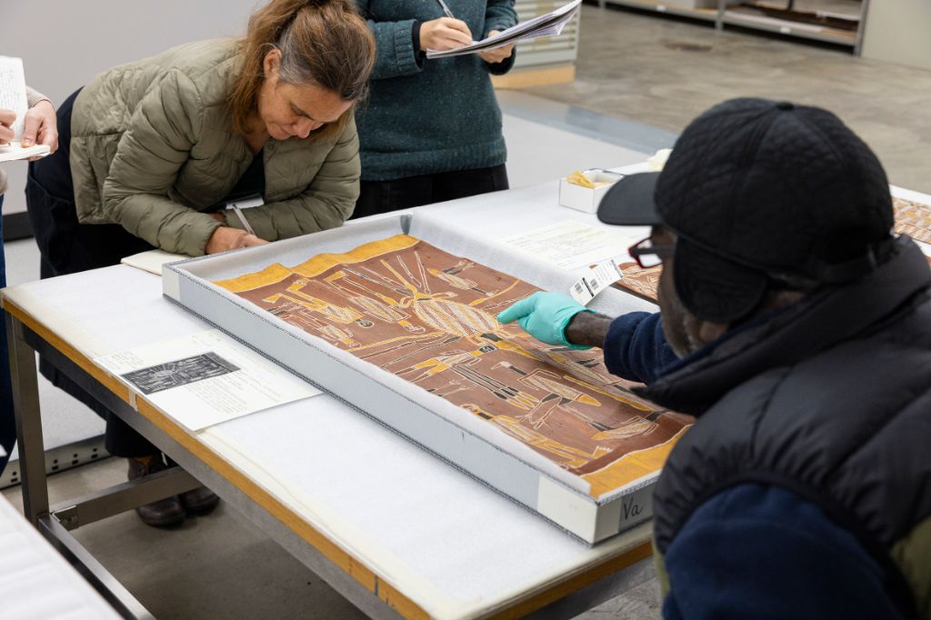 Um einen weissen Tisch sitzen und stehen vier Personen, ein Mann dahinter, drei Frauen davor. Der Mann zeigt auf ein Bild in einer Kartonschachtel mit der rechten Hand, die in einem grünen Handschuh steckt. Die Frauen notieren sich das Gesagt auf Blöcken.