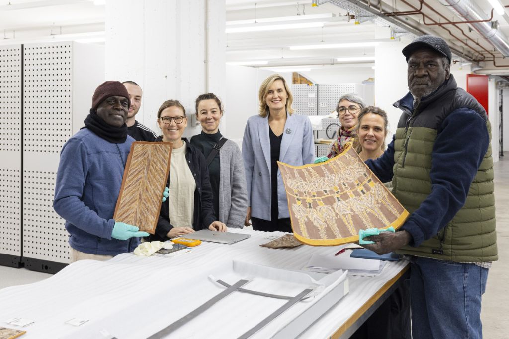 Um den Kopf eines Tisches stehen acht Personen, drei Männer und fünf Frauen. Die Männer halten Bilder in den Händen, die in grünen Handschuhen stecken. Alles lächeln in die Kamera.