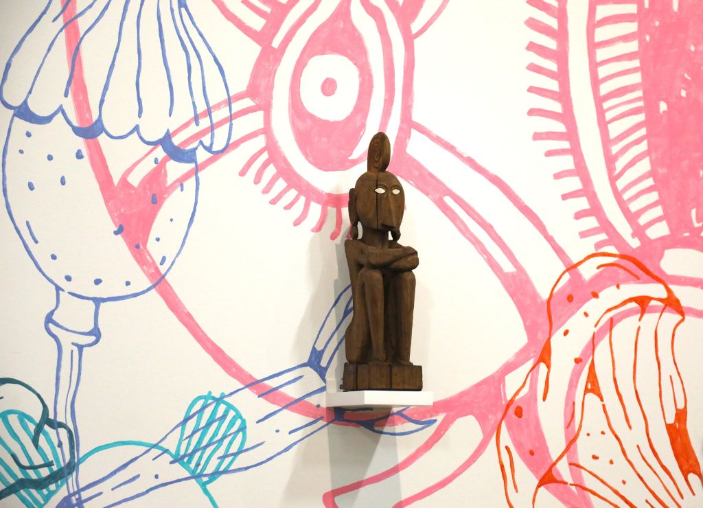 Auf einem weissen Podest an einer weissen Wand, auf der blaue, grüne und rosa Figuren gemalt sind, hockt eine braune Holzfigur. Sie hat ihre Beine angezogen und schaut mit weissen Augen in die Ferne, die Arme hält sie verschränkt auf den Knien.
