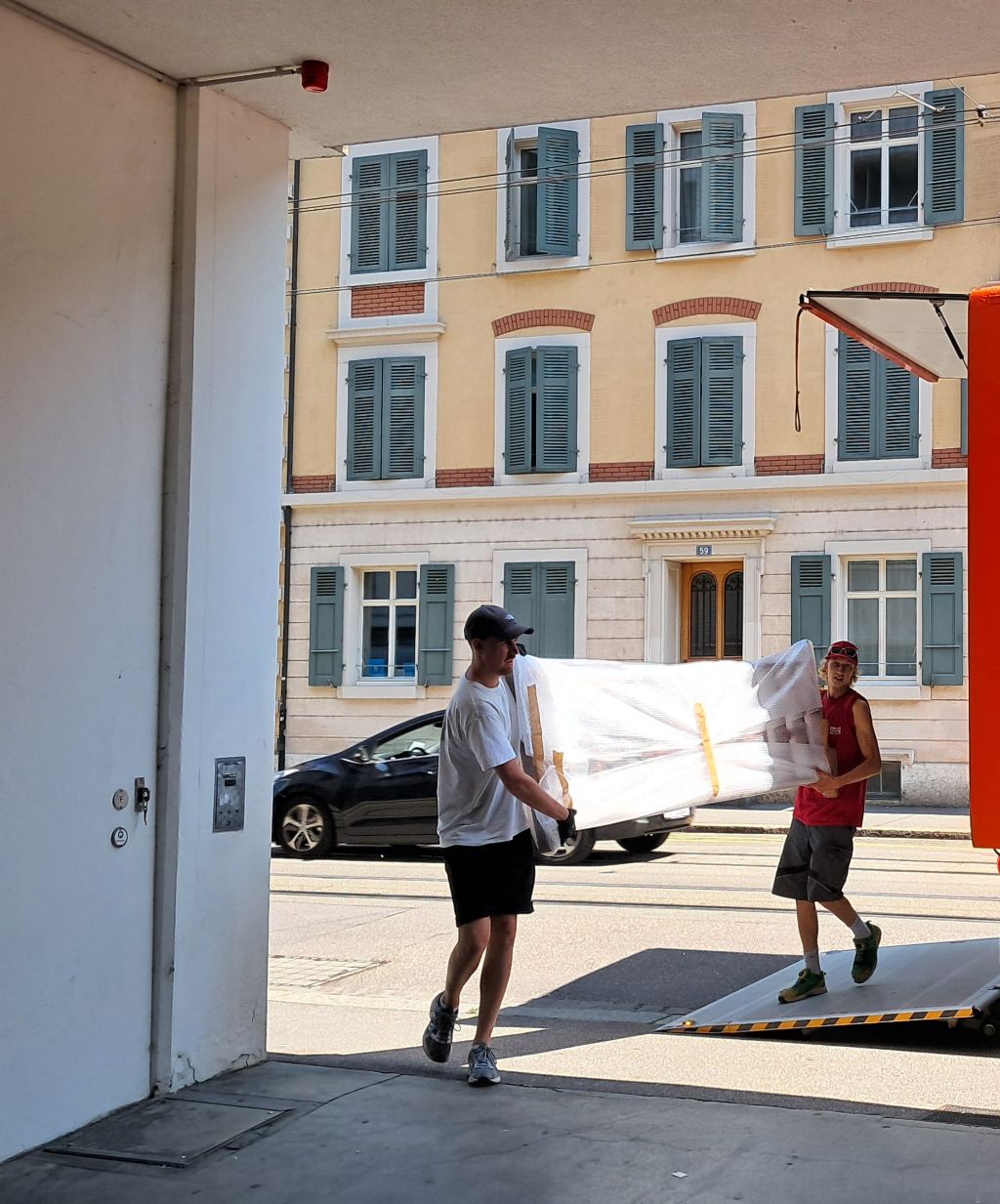 Zwei Männer tragen eine weisseingepackte rechteckige etwa zwei auf einen Meter grosse Platte in ein Gebäude. Im Hintergrund sind eine Strasse mit Tramschienen, ein geparktes Auto und ein mehrstöckiges Haus zu sehen.