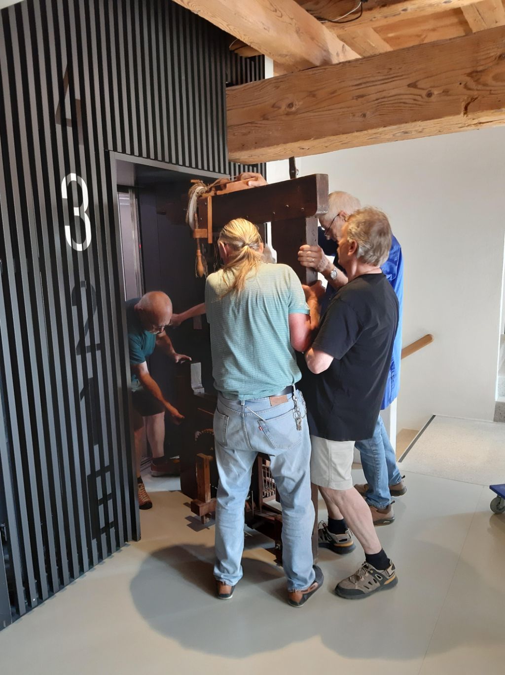 In einem Gebäude mit Holzbalken an der Decke tragen vier Männer ein Gestell aus Holz in einen Lift.
