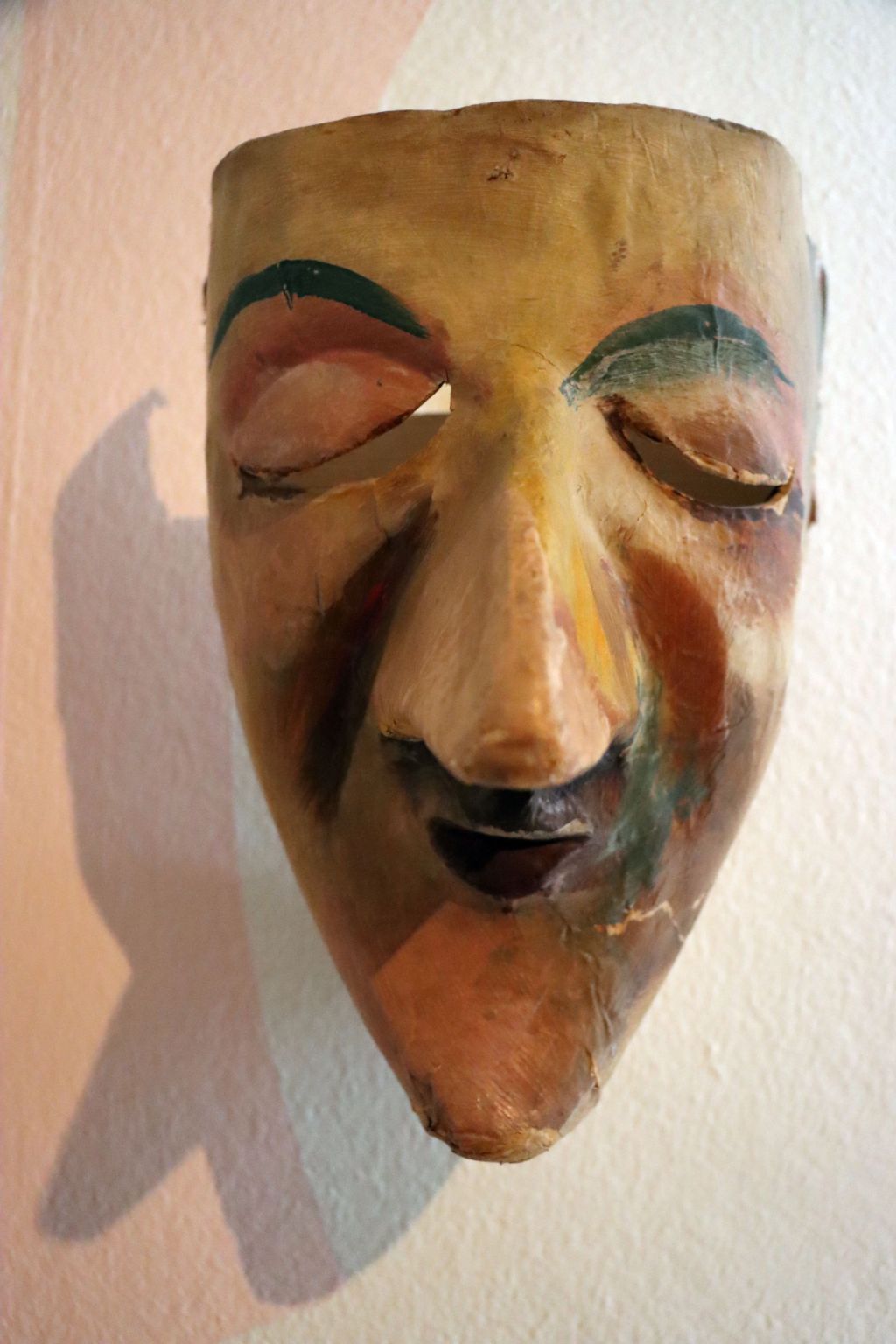 Eine bräunliche, länglich Maske mit geschlossenen Augen, wuchtigen Augenbrauen, einer langen Nase, einem kleinen Mund und einem spitzen Kinn.