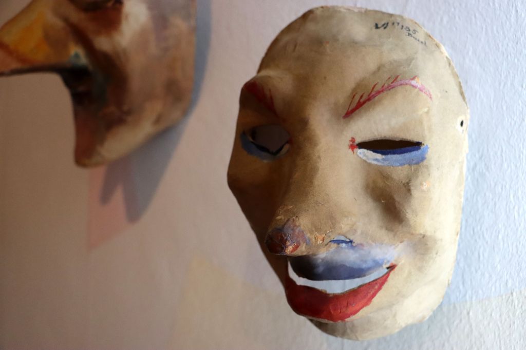 An einer Wand hängen zwei Masken. Die vorne rechts zeigt ein rundes, beiges Gesicht mit Schlitzen als Augen, die unten dran weiss-hellblaue Striche aufweisen. Auch unter der Nase ist die Larve weiss-hellblau bemalt. Der Mund ist gross, die Lippen rot. Die zwei Maske sieht man kaum.