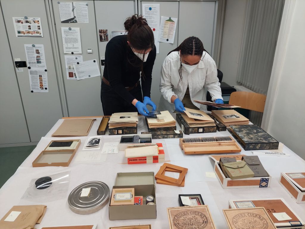 Vor grauen Schränken steht ein grosser Tisch. Darauf sind Fotoalben, einzelne Blätter, Schachteln mit kleinen Schachteln drin und Schachteln mit Fotoblättern drin ausgelegt. Zwei Frauen blättern durch die Fotoblätter aus zwei Schachteln. Sie tragen blauen Handschuhe und Masken.