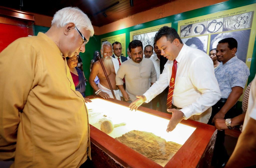 Um eine rote Vitrine scharen sich einige Personen, die auf die Gegenstände schauen, die in der Vitrine ausgestellt sind. Es handelt sich um Objekte aus Stein.