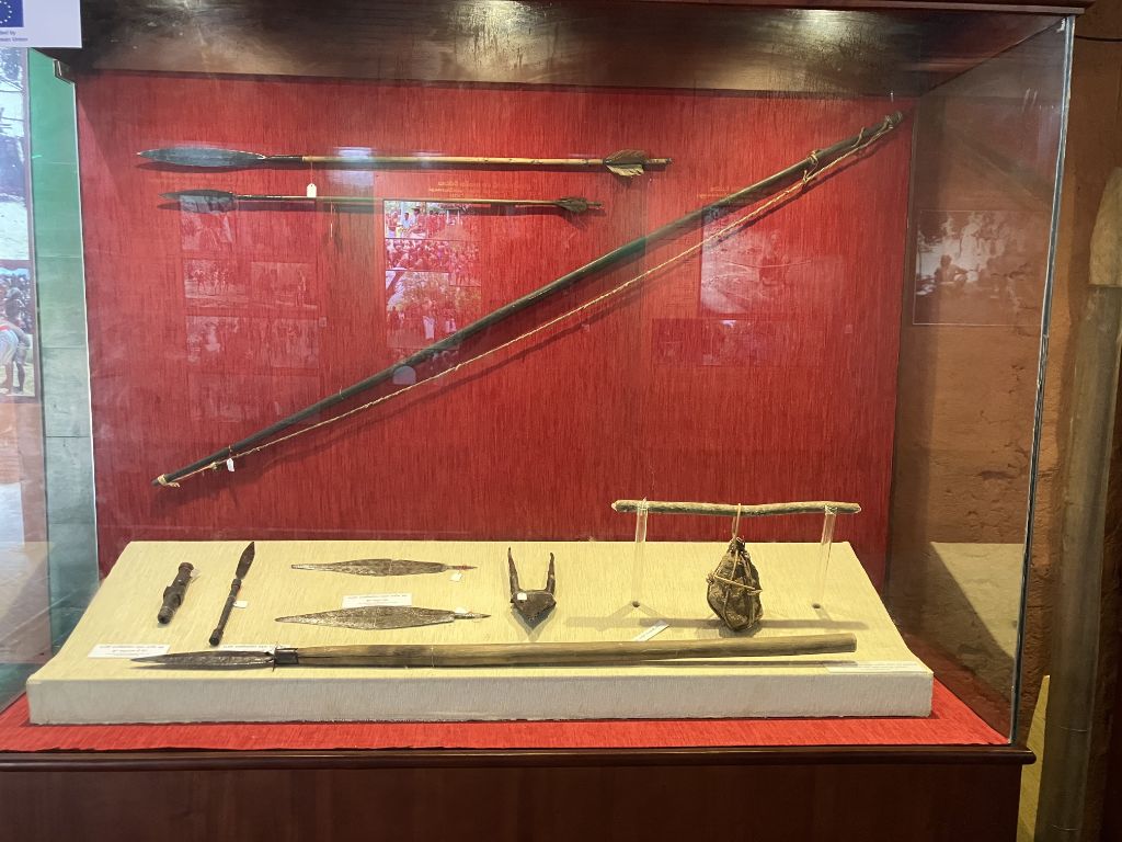 In einer Glasvitrine hängen an einer roten Wand zwei lange Pfeile und ein Pfeilbogen. Auf einer weissen Unterlage liegen sieben Gegenstände aus Holz und Eisen, manche mit Klingen.
