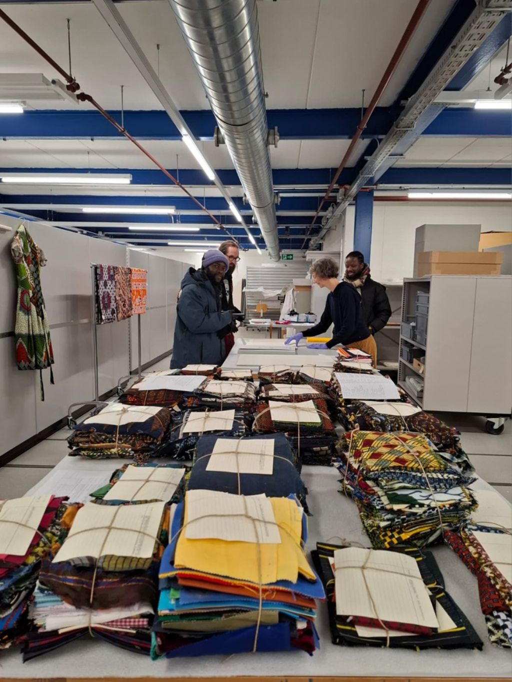 In einer Halle mit silbrigen Röhren an der Decke steht ein sehr langer Tisch und dem viele Bündel Stoff liegen, alle mit Schnur verschnürt und oben drauf jeweils ein Stück Papier. Am einen Ende des Tisches stehen vier Personen.