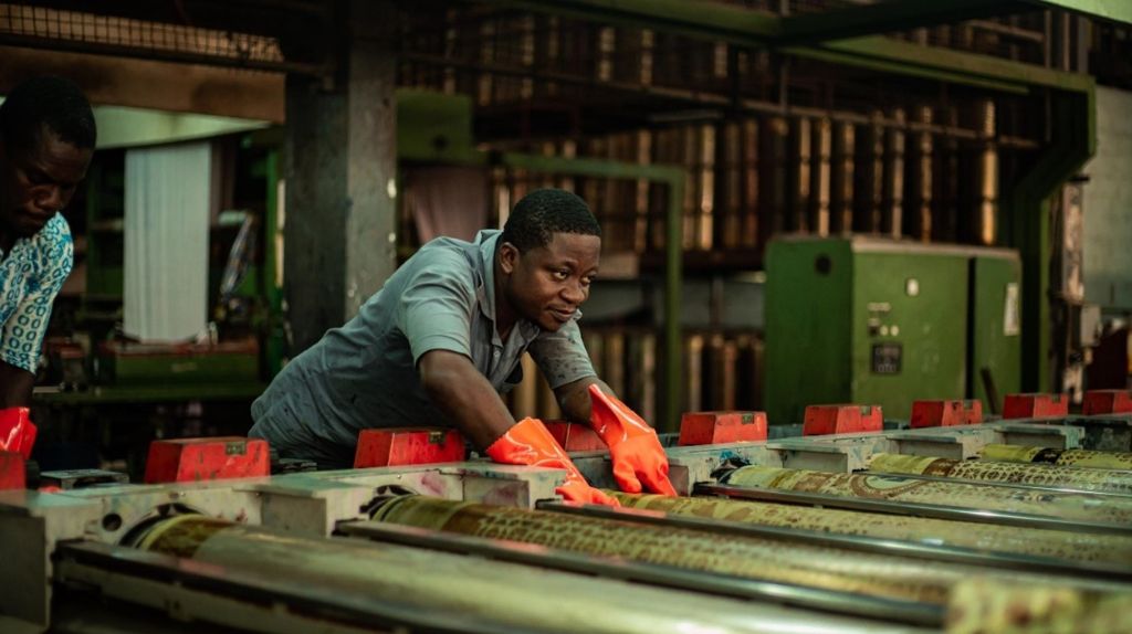 Ein Mann beugt sich über eine Maschine mit vielen Rollen. Er trägt orange Handschuhe und grüne Arbeitskleidung. Hinter ihm sieht man weitere Maschinen.