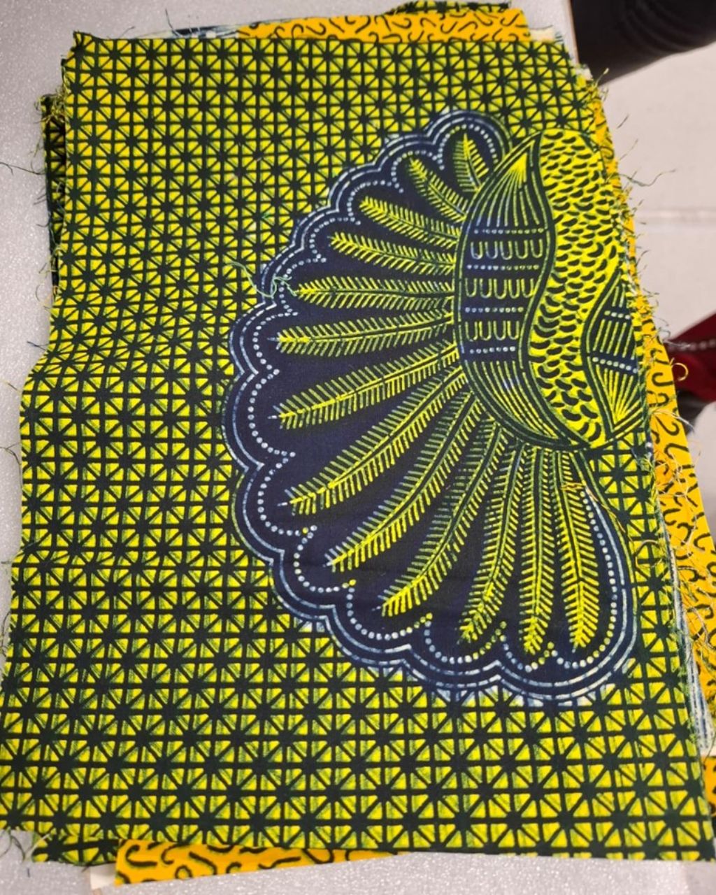 Ein rechteckiges Stück Stoff liegt auf einem Tisch. Es hat ein gelb-blaues Häuschenmuster. Und rechts eine Art Pflanze mit federartigen Blättern.