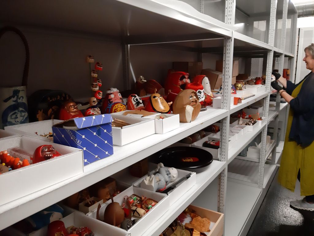 Blick auf eine langes weiss-graues Gestell mit vier Etagen. Auf den unteren Regalen sind einige Schachteln untergebracht, in denen viele kleine, oft handgrosse Figuren liegen und stehen. Auf dem Regal ungefähr auf Augenhöhe stehen viele Figuren auch frei. Ganz am Ende steht eine Frau in olivem Kleide mit schwarzer Jacke, die einen Gegenstand in den Händen hält und leise lächelt.