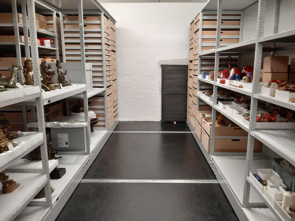 Zwei grau-weisse Rollkorpusse sind auseinandergeschoben. Im linken Gestell sitzen Buddhafiguren und schauen aufs andere Gestell. Im rechten Gestell sind kleine, oft rote Figuren untergebracht, die ihrerseits auf die Buddhas schauen.