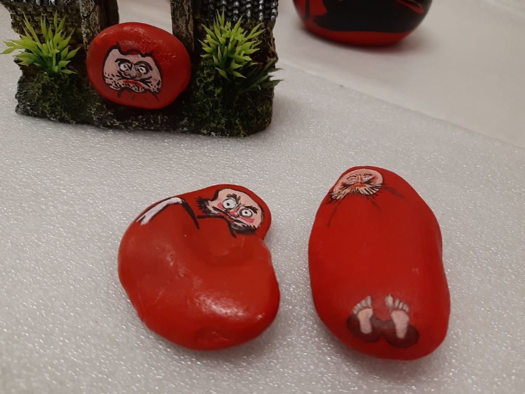 Zwei handflächengrosse längliche Steine liegen auf weisser Unterlage. Sie sind rot bemalt. Der linke Stein weist ein Männergesicht auf mit schwarzem Bart. Der rechte Steine besitzt auch ein aufgemaltes Gesicht und unten aufgemalte Füsse.