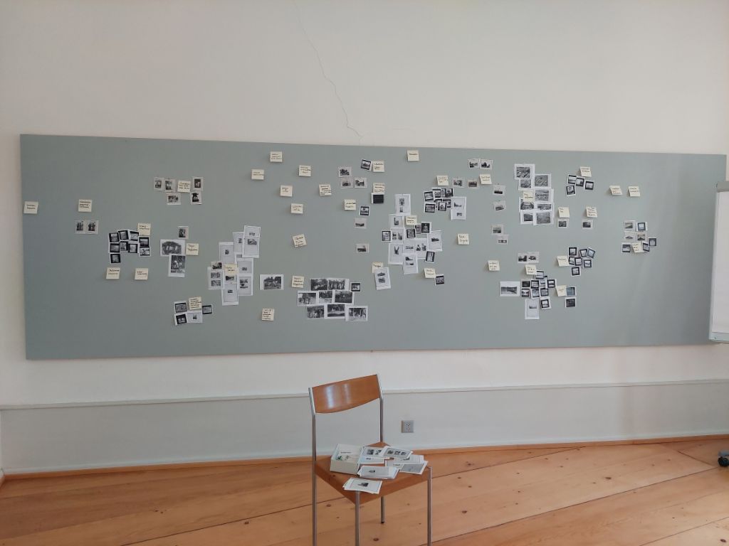 In einem Raum mit Holzboden steht ein Stuhl aus Holz und Metall. Darauf liegen Kopien von Fotos. Dahinter an der weissen Wand hängt eine graue Pinnwand, auf der viele Kopien mehr von Fotos hängen.