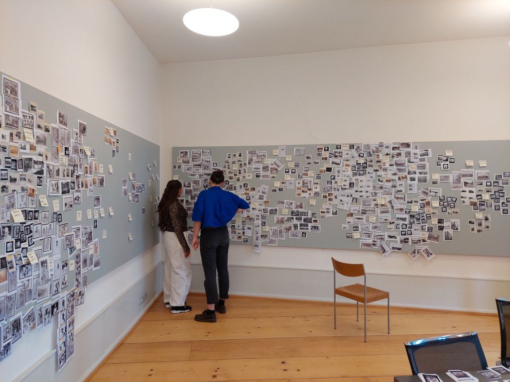 In einem Raum mit Holzboden hängen zwei graue Pinnwände, an denen unzählige Kopien von Fotos hängen. Zwischen den Pinnwänden in einer Ecke stehen zwei Personen, die Fotos betrachten. 