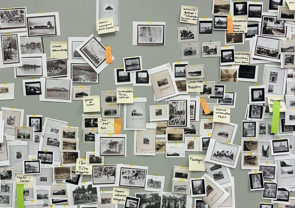 An einer grauen Pinnwand hängen in Reihen Kopien von Schwarz-Weiss-Fotos. Auf einigen Fotos sind orange oder grüne längliche Post-its angebracht.