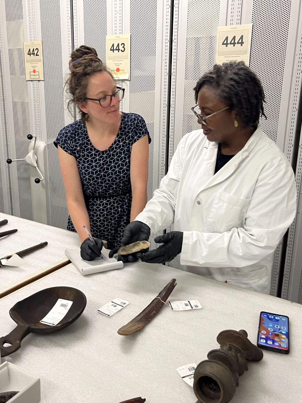 Vor Rollgestellen steht ein weisser Tisch, an dem zwei Frauen stehen. Sie tragen schwarze Handschuhe und die eine Frau hält einen Gegenstand in den Händen, über den sie offensichtlich sprechen. Vor ihnen auf dem Tisch liegen noch einige Objekte aus Holz und ein Natel.