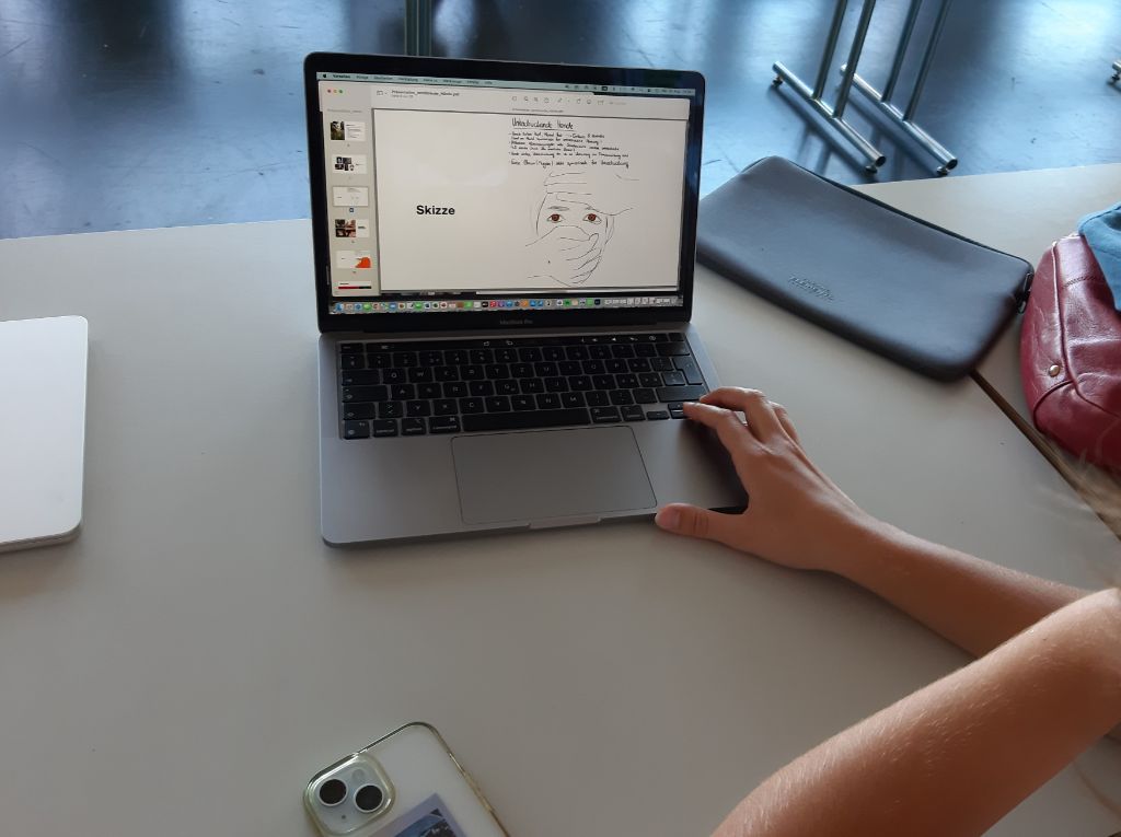 Auf einem Laptop ist eine Skizze eines Kopfes einer Frau zu sehen, den zwei Hände an der Stirn und über den Mund umfassen. Eine Hand berührt die Tastatur des Laptops, der auf einem grauen Tisch steht.