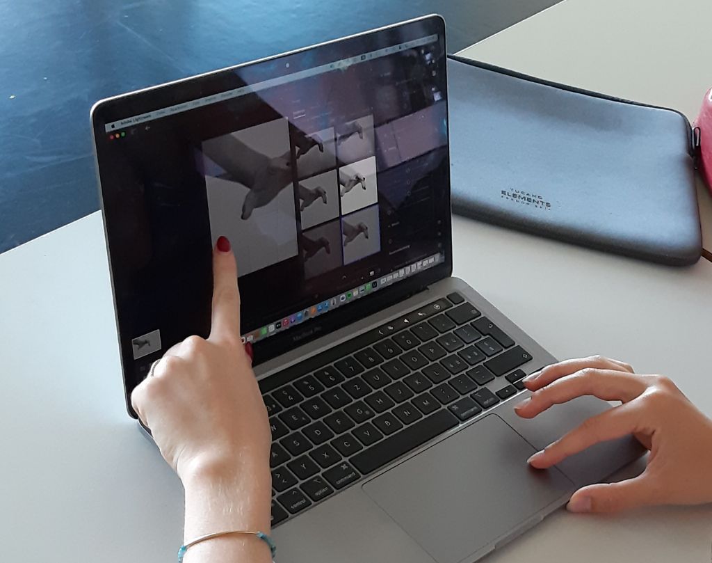 Auf einem grauen Laptop sind auf dem Bildschirm Schwarz-Weiss-Fotos von Händen zu sehen. Eine Hand mit rot angemalten Fingernägeln weist auf die Fotos. Eine andere Hand berührt mit den Fingern den Laptop.