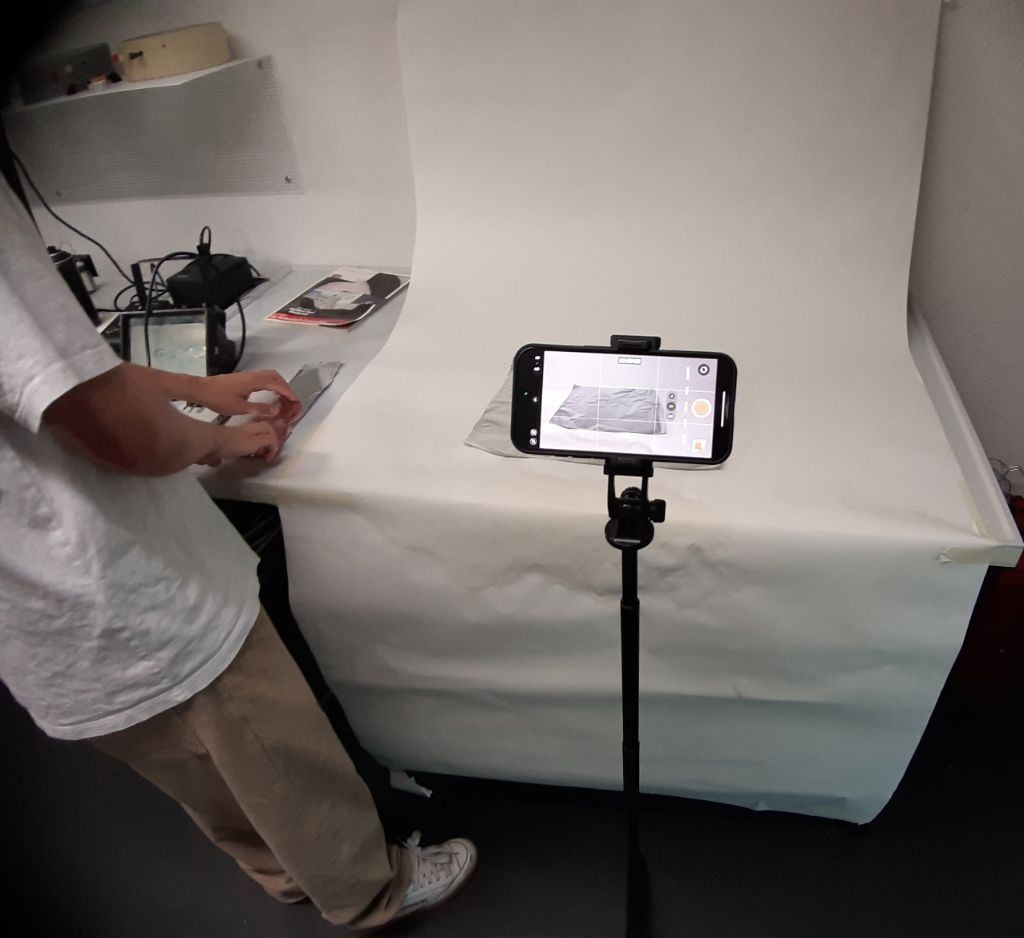 Vor einem Tisch, auf den ein weisses Papier gespannt ist, bis an die Wand, steht ein Ständer mit einem Handy drauf befestigt. Auf dem Handy sieht man eine Alufolie, die auf dem Tisch liegt. Links sind die Beine, der Oberkörper und die Hände einer Person zu sehen, die etwas auf dem Tisch berührt. Die Person trägt ein weisses T-Shirt und beige Hosen.