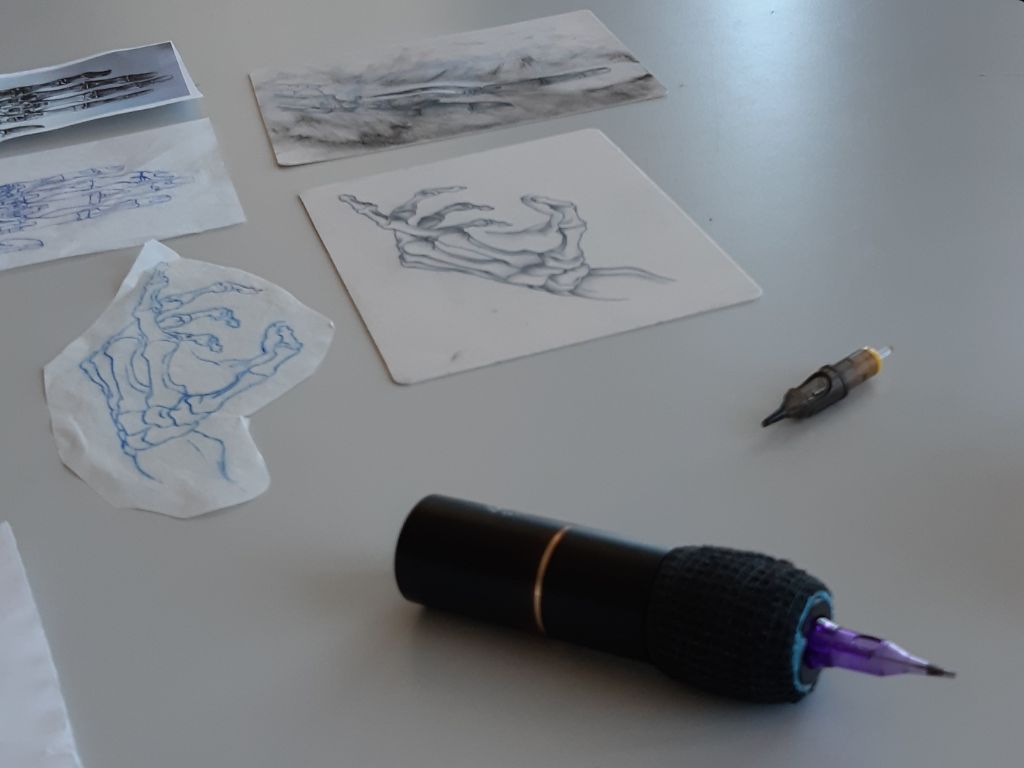 Auf einem grauen Tisch liegt im Vordergrund ein schwarzer Tätowierstift mit einer lila Feder. Dahinter liegen eine zweite Feder und fünf Zeichnungen von Händen.