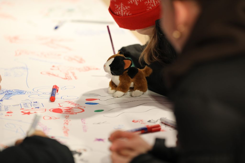 Menschen, die man nicht erkennt, schreiben auf ein grosses weisses Papier, auf dem schon viel steht. Auf dem Papier steht zudem ein kleines Plüschkätzchen.