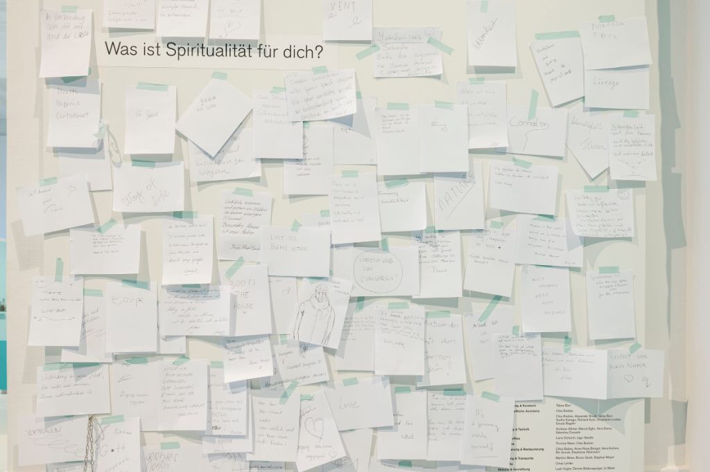 Weisse Wand, an der viele kleine weisse Zettel hängen, die mit blauem Kugelschreiber beschriftet sind. In schwarzer Schrift steht links oben auf der Wand: Was ist Spiritualität für dich?
