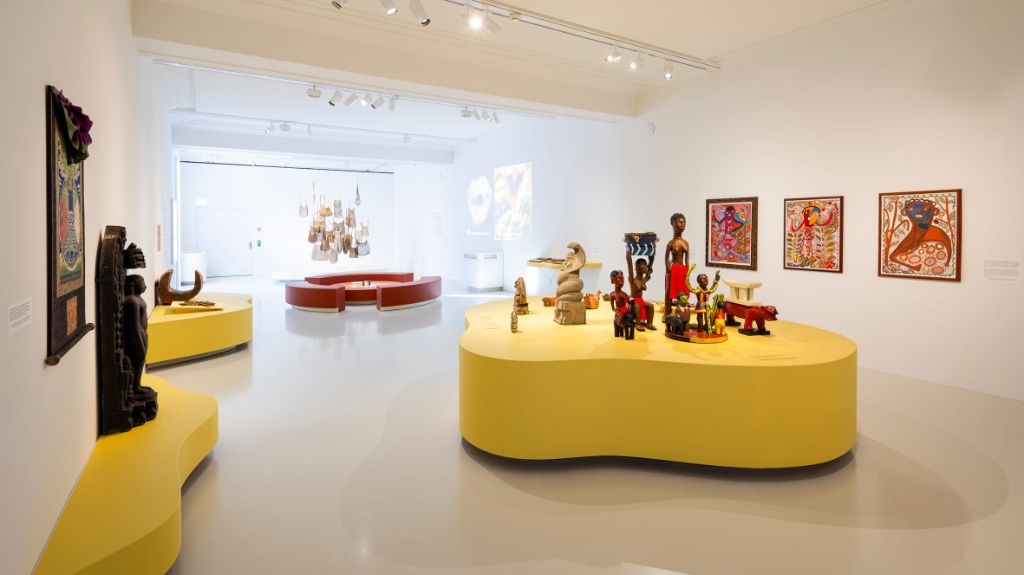 Ein heller Ausstellungsraum mit farbenfrohen Skulpturen und Gemälden auf gelben Podesten