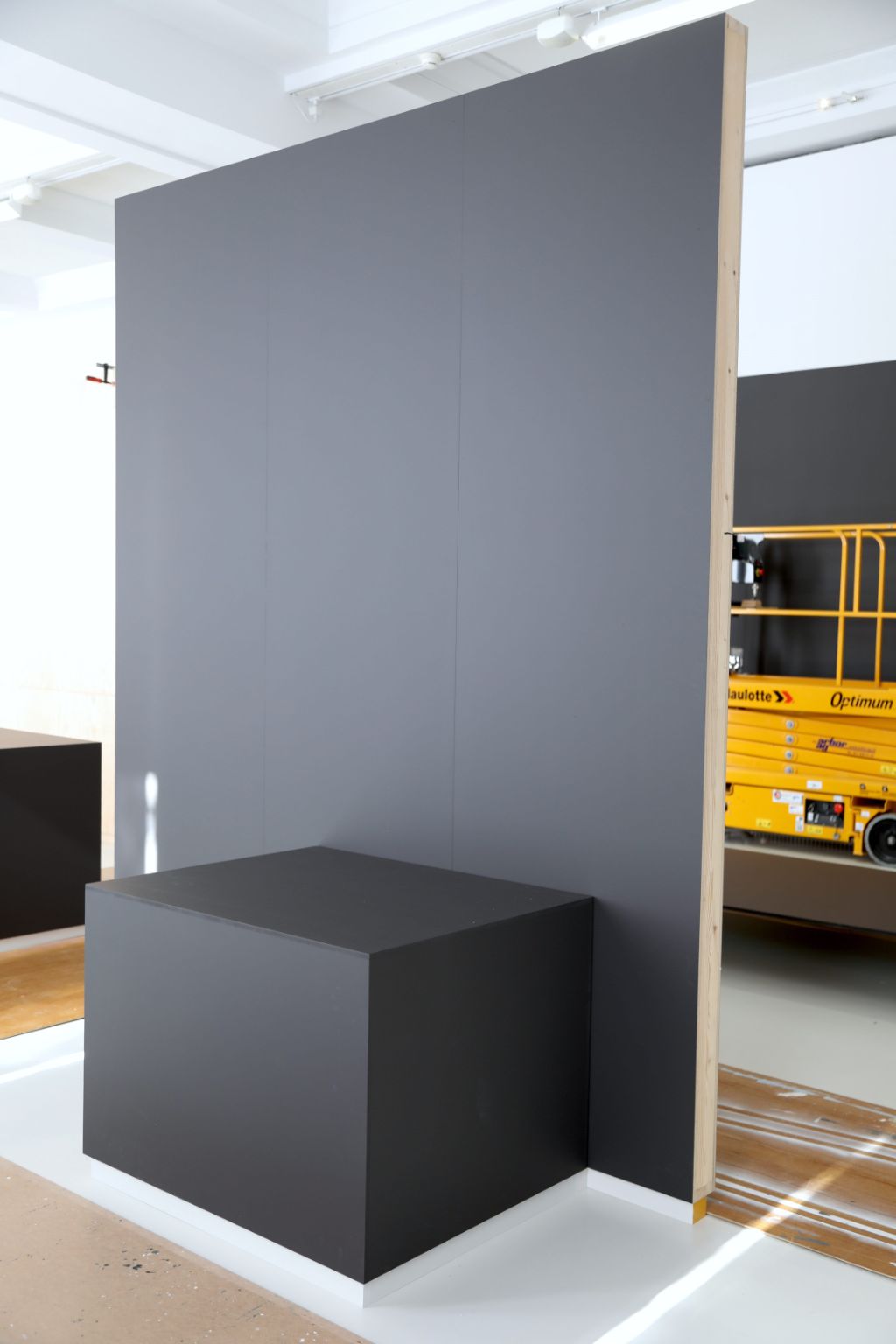 Ein grosser grauer Kubus mit Holzkanten steht in einem hellen Raum vor gelber Hebebühne
