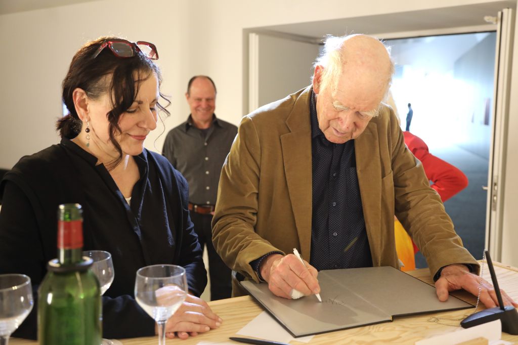 Eine Frau lächelt, während ein älterer Mann in ein Buch signiert. Im Hintergrund sind zwei Personen sichtbar.