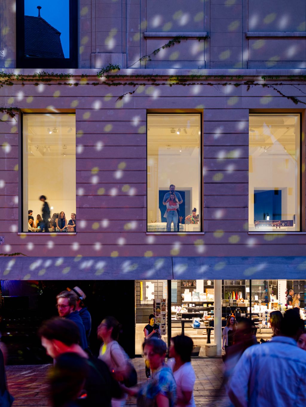 Menschen gehen vor einem beleuchteten Gebäude mit grossen Fenstern und schimmernden Lichtern vorbei