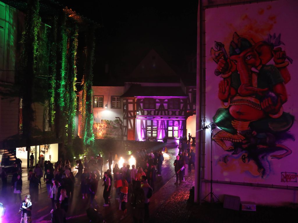 Eine Gruppe von Menschen feiert nachts draussen, umgeben von farbenfrohen Lichtern und Wandgemälde