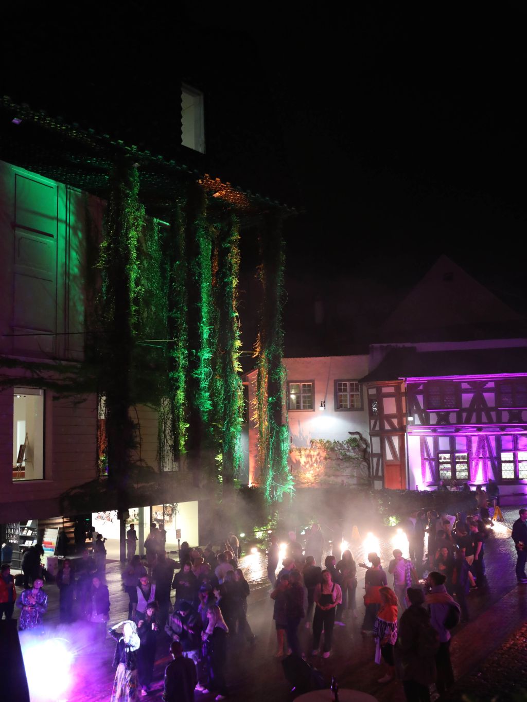 Auf einem nächtlichen Platz tanzen Menschen unter buntem Lichtschein vor alten Gemäuern