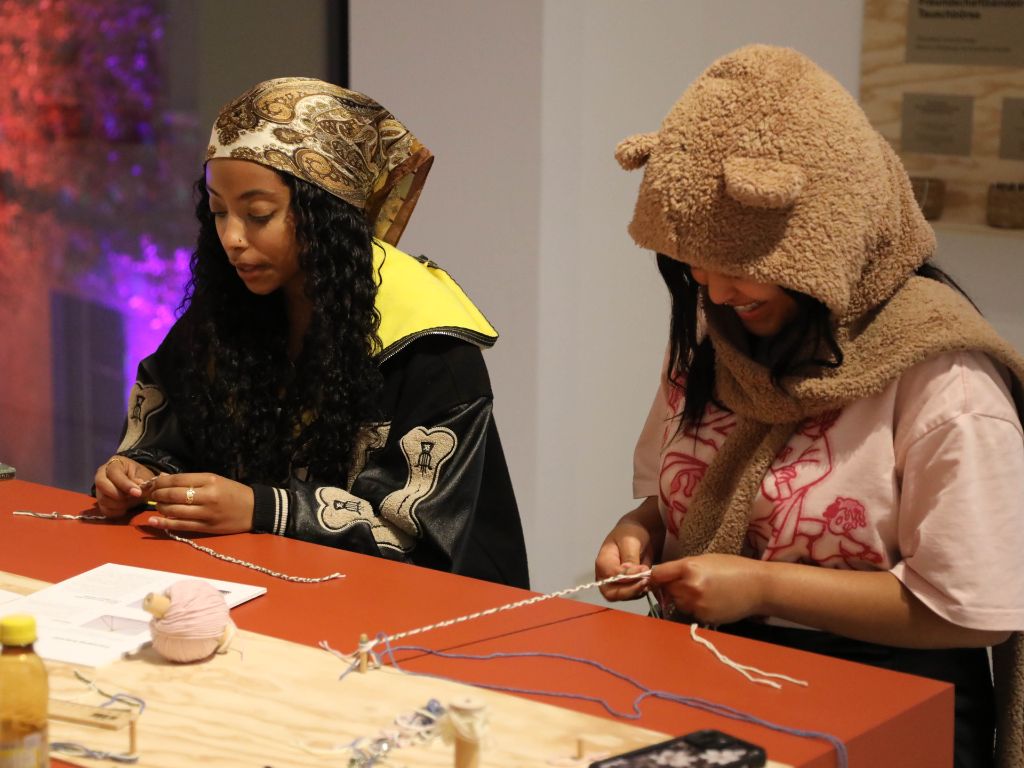 Zwei Frauen sitzen an einem Tisch und flechten bunte Schnüre