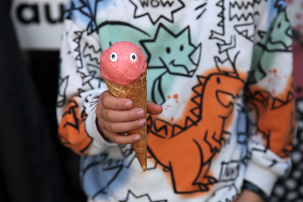 Kinderhand hält ein Eis in der Waffel. Die Eiskugel ist rosarot und hat zwei Zuckeraugen. Das Kind trägt ein langärmliges T-Shirt mit bunten Dinosauriern.