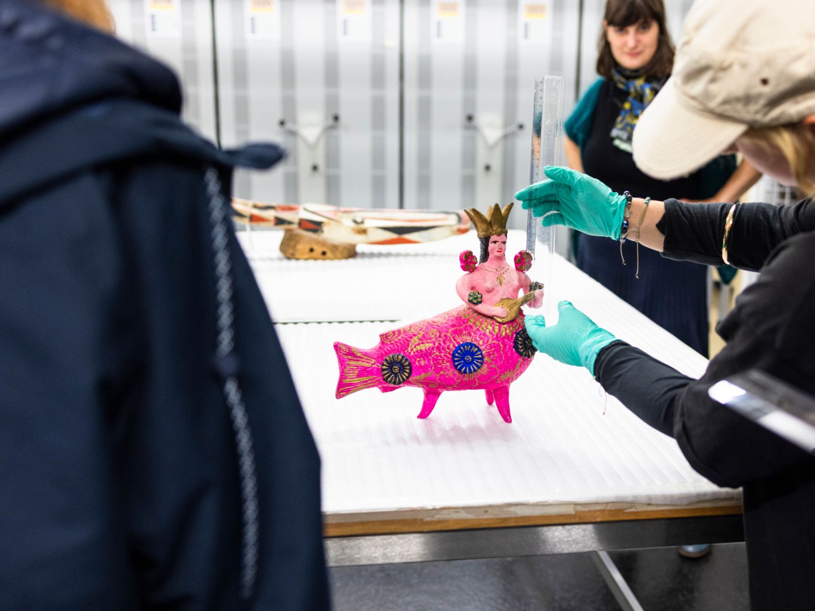 Auf einem weissen Tisch steht die Figur einer Meerjungfrau mit dickem pinkem Fischschwanz, nacktem Oberkörper und einer Krone auf Kopf. Links die Umrisse einer Person von hinten, rechts von der Figur Arme und Hände in blauen Handschuhen einer Person, und weiter im Hintergrund eine weitere  Person.